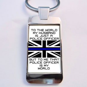 May include: A silver keychain with a white background and black text that reads "To the world my husband is just a police officer, but to me that police officer is my world". The background also features a black and white Union Jack flag with a blue stripe across the middle.