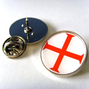 May include: A silver pin with a red cross on a white background. The pin has a safety clasp on the back.