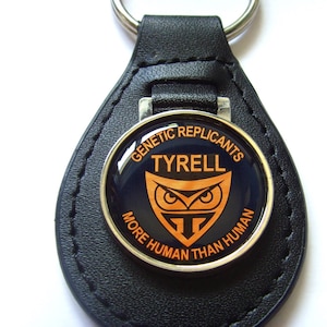 May include: Black leather keychain with a silver round charm featuring an orange owl logo and the text "Genetic Replicants Tyrell More Human Than Human".