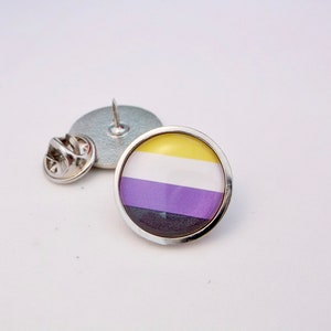 Pode incluir: Um alfinete de prata com um design redondo e colorido. O alfinete apresenta um padrão listrado amarelo, branco, roxo e preto, representando a bandeira do orgulho não binário.
