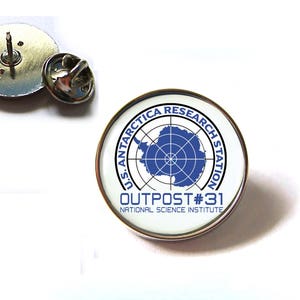Op de afbeelding: Een witte emaille pin met een blauw en wit ontwerp. De pin heeft een cirkel met de tekst "U.S. ANTARCTICA RESEARCH STATION" rond de buitenkant. Binnen de cirkel bevindt zich een blauwe kaart van Antarctica met een radar-achtig ontwerp. De tekst "OUTPOST #31" staat onder de cirkel, en de tekst "NATIONAL SCIENCE INSTITUTE" staat daaronder.