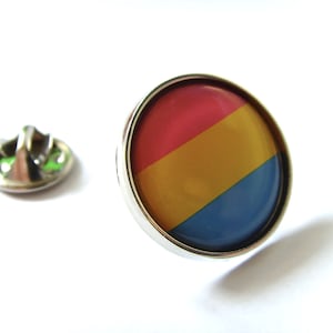 Puede incluir: Un pin redondo con un diseño rojo, amarillo y azul. El pin está hecho de metal y tiene un acabado plateado.
