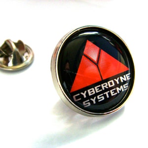 Könnte beinhalten: Eine silberne Anstecknadel mit einem schwarzen und roten Logo. Das Logo ist ein rotes Dreieck mit weißem Rand und dem Text "CYBERDYNE SYSTEMS" in weißen Buchstaben.