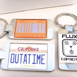 Könnte beinhalten: Drei silberne Schlüsselanhänger mit unterschiedlichen Designs. Der erste Schlüsselanhänger hat ein orangefarbenes und violettes Nummernschild mit dem Text "CALIFORNIA OUTATIME The Golden State". Der zweite Schlüsselanhänger hat ein orangefarbenes und violettes Nummernschild mit dem Text "CALIFORNIA" und einem Barcode. Der dritte Schlüsselanhänger hat einen weißen Hintergrund mit dem Text "FLUX CAPACITOR" und einem stilisierten Bild eines Flux-Kondensators.