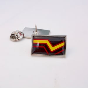 Può includere: Uno spillo d'argento con un designo a strisce nere, rosse e gialle. Lo spillo è rettangolare e ha un retro in argento.