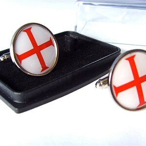 May include: A pair of silver cufflinks with a white background and a red cross design. The cufflinks are in a black box.