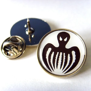 Op de afbeelding: Een witte emaille pin met een gouden metalen frame. De pin heeft een zwart-witte afbeelding van een gestileerde octopus met een dreigende uitdrukking.