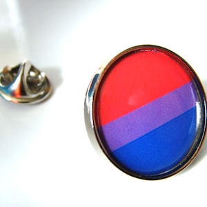 Puede incluir: Un pin de plata con un diseño redondo de esmalte rojo, morado y azul. El pin es una representación de la bandera del orgullo bisexual.