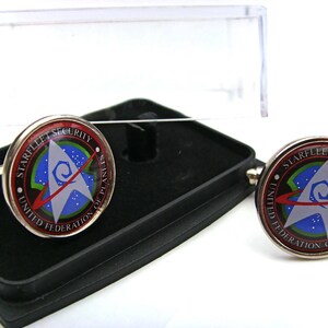 Puede incluir: Un par de gemelos de plata con un logotipo de Starfleet Security rojo, verde y azul. El logotipo presenta una nave espacial estilizada y el texto "Starfleet Security United Federation of Planets".