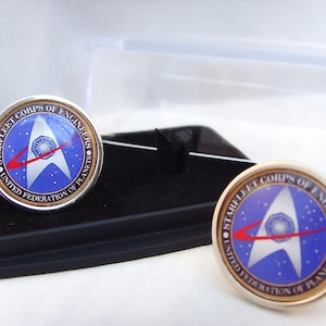 May include: A pair of silver cufflinks featuring a blue and gold Starfleet Corps of Engineers emblem. The emblem depicts a white Starfleet delta with a red arc and a white star at its centre. The text surrounding the emblem reads "Starfleet Corps of Engineers United Federation of Planets".