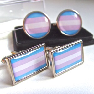 Puede incluir: Un par de gemelos de plata con un diseño de bandera del orgullo transgénero. La bandera es azul claro, rosa y blanca.