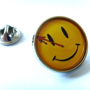 Peut inclure: Une épingle en forme de smiley jaune avec une barre rouge sur l'œil. L'épingle est en métal et a une finition argentée.