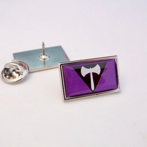 Può includere: Uno spillo in smalto viola con bordo argentato e un simbolo bianco di un'ascia in un triangolo. Lo spillo ha un retro argentato con una spilla di sicurezza.
