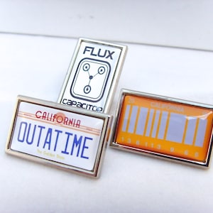 以下が含まれることがあります： 異なるデザインの銀色のピンが3つあります。最初のピンは、白い正方形で、「FLUX CAPACITOR」というテキストと、回路基板のスタイリッシュな画像が描かれています。2番目のピンは、白い長方形で、「CALIFORNIA OUTATIME」というテキストと、赤と青のデザインが描かれています。3番目のピンは、黄色の長方形で、「CALIFORNIA」というテキストと、数字のバーコードが描かれています。