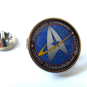 Puede incluir: Un pin de plata con un logotipo de Starfleet Command azul, blanco y amarillo. El logotipo presenta una nave espacial estilizada y el texto "Starfleet Command United Federation of Planets".