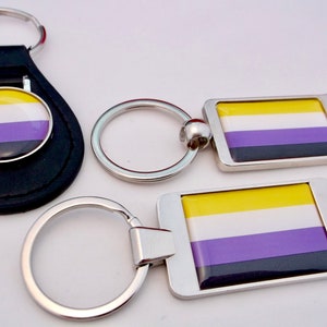Peut inclure: Trois porte-clés différents représentant les couleurs jaune, blanc, violet et noir du drapeau de la fierté non binaire. Un porte-clés est un porte-clés en cuir noir avec un charme rond en métal, un autre est un porte-clés argenté avec un charme rectangulaire en métal, et le troisième est un porte-clés argenté avec un charme rectangulaire en métal et un ouvre-bouteille.