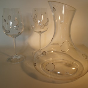 Puede incluir: Una jarra de vino de cristal transparente y dos copas de vino. Cada pieza está decorada con un patrón de óvalos grises. La jarra tiene una base ancha y un cuello delgado. Las copas tienen tallos largos y cuencos redondeados.