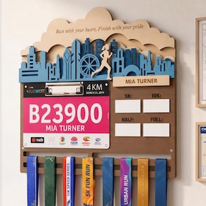 May include: A wooden wall-mounted race medal and bib display rack featuring a layered city skyline silhouette, a metal clip for a race bib with the text B23900 MIA TURNER, and eight hooks holding various colorful race medals with ribbons.
