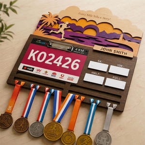 May include: A dark wood medal display rack featuring a layered sunset scene with a palm tree and runner silhouette. It includes a metal clip holding a race bib numbered K02426, a nameplate for John Smith, and slots for 5K, 10K, half, and full marathon times.