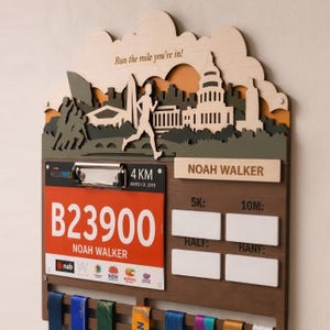 May include: A wooden race medal display board featuring a layered skyline silhouette, a runner figure, and the text Run the mile you're in. It includes a metal clip holding a race bib for Noah Walker, 4 KM, and blank white spaces for 5K, 10M, Half, and Hanf.