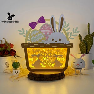 Pode incluir: Uma luz noturna de coelho da Páscoa de madeira com uma cesta cheia de ovos coloridos. O coelho é branco com orelhas cor-de-rosa e um laço roxo. A cesta é marrom com um interior branco. A luz noturna é iluminada com um brilho amarelo quente.