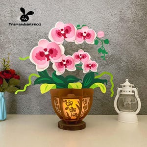Può includere: Una composizione di orchidee di carta con fiori rosa e bianchi, foglie verdi e un vaso marrone con un design ritagliato. Il vaso poggia su una base di legno e una piccola luce illumina l'interno.