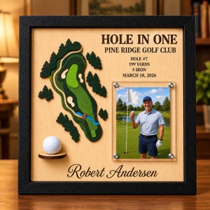 May include: A framed wooden display featuring a 3D layered map of a golf course, a white golf ball on a small shelf, and a photograph of a person on a green. Text reads: HOLE IN ONE, PINE RIDGE GOLF CLUB, HOLE #7, 199 YARDS, 5 IRON, MARCH 18, 2026, Robert Andersen.