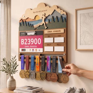 May include: A wooden wall-mounted medal display rack featuring a layered mountain landscape with a runner silhouette and the text Run the mile you're in. It includes a clipboard for a race bib, slots for race times, and eight hanging race medals.