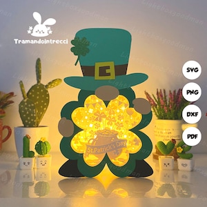 Puede incluir: Lámpara recortada de papel con temática del Día de San Patricio con forma de duende. La lámpara presenta un sombrero verde con hebilla dorada, un trébol de cuatro hojas y un diseño de trébol brillante. El texto "St. Patrick's Day" es visible.