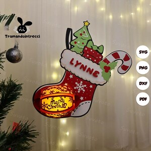 Può includere: Decorazione a calza natalizia rossa e bianca con il nome "LYNNE". La calza presenta un albero di Natale, un regalo e un bastoncino di zucchero. Una sfera luminosa con "Merry Christmas" è attaccata in basso.