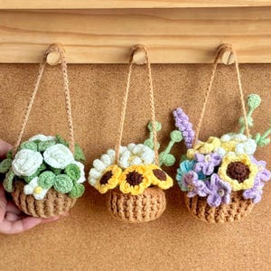 Può includere: Tre cestini di fiori all'uncinetto fatti a mano. Ogni cestino marrone ha una trama intrecciata e un manico in corda intrecciata. I fiori sono bianchi, gialli e viola, con foglie verdi. I cestini sono appesi a pioli di legno.