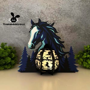 Può includere: Una lanterna decorativa a tema cavallo con manico nero e design traforato. La lanterna presenta una silhouette di cavallo, montagne e alberi. Lo sfondo include una testa di cavallo e alberi. Il testo "Tramandointrecci" è visibile.