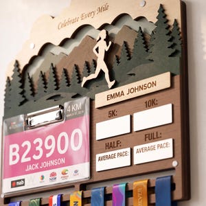 May include: A wooden wall-mounted display for race medals and bibs featuring a layered mountain and forest scene with a runner silhouette. It includes the text "Celebrate Every Mile," "Emma Johnson," and "Jack Johnson" on a race bib, with slots for 5K, 10K, Half, and Full marathon times.