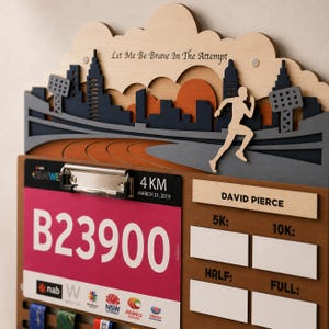 May include: A wooden race bib display board featuring a layered city skyline, a running figure, and the text Let Me Be Brave In The Attempt. It includes a metal clip holding a pink race bib numbered B23900, with slots for medals and blank spaces for race times.