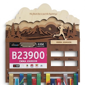 May include: A wooden medal display rack featuring a layered mountain and forest landscape with a runner silhouette. It includes a clipboard holding a pink race bib for Emma Johnson, a name plate, and four slots labeled 5K, 10K, HALF, and FULL.