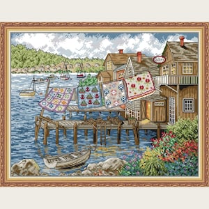 Könnte beinhalten: Ein gerahmtes Kreuzstichbild, das eine Küstenlandschaft mit einem Holzsteg, Booten und Gebäuden zeigt. Bunte Quilts hängen an einer Wäscheleine. Das Wasser ist blau und der Himmel ist bewölkt. Das Wort "Quilts" steht auf einem Schild.