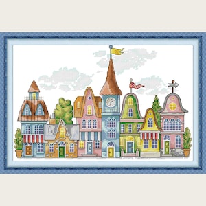 Può includere: Un punto croce di una scena cittadina colorata con vari edifici. Gli edifici hanno diversi stili architettonici e colori, tra cui blu, rosa, giallo e verde. Sono visibili una torre dell'orologio e delle bandiere. L'opera d'arte è incorniciata in un bordo blu.
