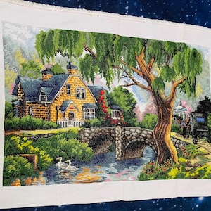May include: A completed cross-stitch landscape featuring a stone bridge, a house with a dark blue roof, and a large tree. The scene includes a river with two white swans, a horse-drawn carriage, and lush greenery. The artwork is on white fabric.