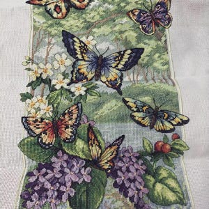 May include: A detailed needlepoint depicting butterflies in shades of orange, yellow, blue, and purple. The butterflies are arranged around a floral display with purple flowers and green foliage, set against a backdrop of a forest scene.