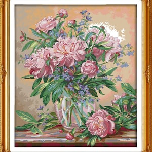 May include: A framed cross-stitch floral arrangement featuring a bouquet of pink peonies, blue wildflowers, and green leaves in a clear glass vase. The artwork is set against a neutral background and framed in a gold ornate frame.