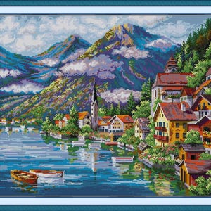 May include: Cross-stitch landscape of a lakeside village nestled at the foot of majestic mountains. The scene features colorful houses with red roofs, a church steeple, and boats on the water. The sky is filled with clouds.