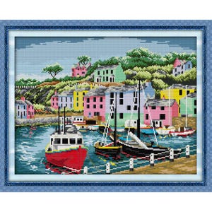 May include: A framed cross-stitch depicting a colorful harbor scene. The artwork features a red boat, sailboats, and buildings in shades of pink, yellow, blue, and green. The water is a deep blue, and the sky is light blue.