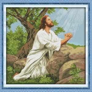 May include: A cross-stitch depiction of a man with long brown hair and a beard, dressed in white robes, kneeling in prayer. The scene is set outdoors, with a tree, rocks, and sunlight streaming down from above. The image is framed with a blue border.