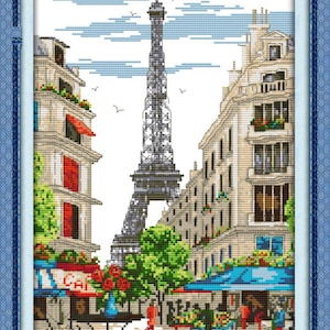 May include: Cross-stitch artwork depicting a Parisian street scene with the Eiffel Tower in the background. Buildings with red awnings and flower boxes line the street. A cafe with the word "Cafe" is visible. The artwork is framed in a blue border.