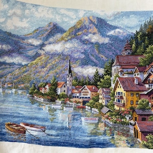 May include: Cross-stitch landscape showing a lakeside village set against towering mountains. The image features colorful houses, a church with a tall spire, and two small boats on the water. The mountains are a blend of blues, greens, and purples.
