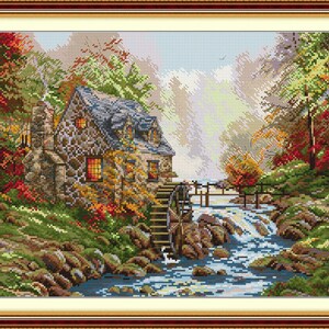 May include: Cross-stitch artwork depicting a stone cottage with a water wheel beside a flowing stream. The scene is framed by autumn foliage in shades of red, orange, and green, with a wooden bridge in the background. The artwork is in a decorative wooden frame.