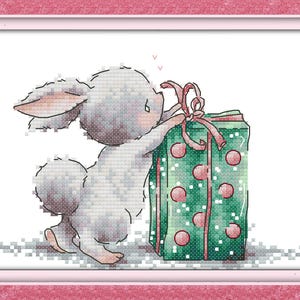 May include: A white bunny with gray ears and a fluffy tail is holding a green gift box with red polka dots and a red ribbon.