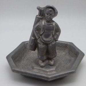 Può includere: Una statuetta in metallo color argento di un golfista che tiene una sacca da golf con una mazza che sporge. La statuetta è in piedi su un vassoio esagonale.