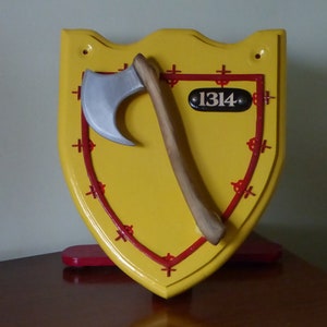 May include: A yellow wooden shield with a red border and a silver axe attached. The shield has a black oval with the number 1314 on it.