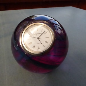 Puede incluir: Un pisapapeles de vidrio morado con una esfera de reloj plateada que dice "Caithness Crystal Quartz".